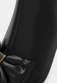 Zwarte leren schoen met een gladde afwerking, voorzien van een grote satijnen strik en een dunne gouden accent waar de strik is bevestigd.