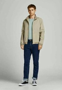 Young man standing wearing a beige zip-up jacket, light blue shirt, dark blue rolled-up jeans, and black sneakers with white laces.