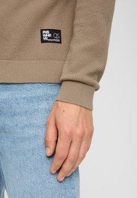 Brauner strukturierter Pullover mit geripptem Saum und Bündchen, getragen über blauer Jeans. Besitzt ein schwarzes Etikett mit weißem Text am Ärmel.
