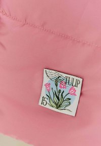 Ružová polstrovaná tkanina so šitým priestorom, ktorý zobrazuje štvorcovú nálepku s tulipánmi a textom "TULIP" a "15." Hladká štruktúra.