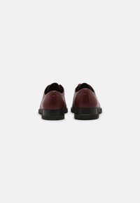 Paire de chaussures habillées en cuir bordeaux vues de l'arrière avec des semelles noires sur un fond blanc uni.
