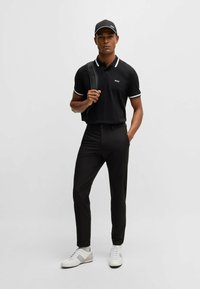 Polo nero con accenti bianchi, pantaloni neri e scarpe da ginnastica bianche. Il modello indossa uno zaino nero e un cappellino nero.