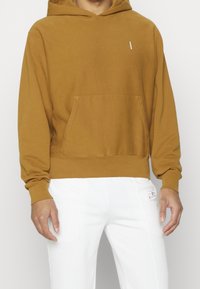 Felpa marrone con cappuccio, tasca frontale, polsini e orlo a costine. Cappuccio regolabile, piccolo logo sul petto, indossata con pantaloni bianchi. Tessuto morbido.