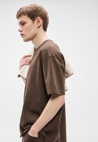 Brązowy T-shirt z krótkim rękawem o luźnym fasonie, o gładkiej fakturze. Jasnokolorowy sweter z dzianiny zarzucony na ramię. Prosty wzór.