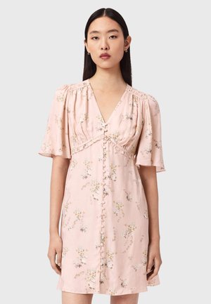 Kobieta ubrana w jasnoróżową sukienkę w kwiaty z falbanowymi rękawami i zapinana z przodu na guziki, stoi na tle jednolitego jasnoszarego tła.