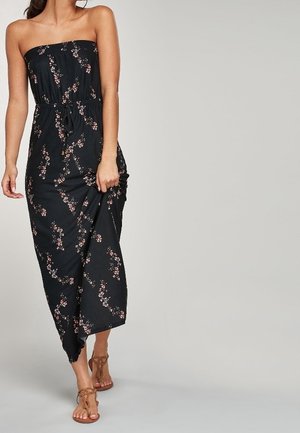 Robe maxi noire sans manches avec un motif floral, taille froncée et tissu fluide. Portée avec des sandales à talons beige. Présente une silhouette décontractée.