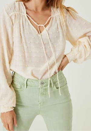 Nainen pukeutuneena teksturoituun kermaiseen paitaan, jossa on solmittava kaula-aukko ja ilmapallohihat, yhdistettynä vaaleanvihreisiin korkeavyötäröisiin farkkuihin valkoista taustaa vasten.