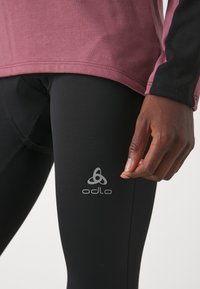 Fekete leggings, az Odlo logójával a combon, egy laza, bordó és fekete, hosszú ujjú felsővel párosítva, amely sima szövet-textúrát mutat.
