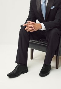 Schwarze Lederschuhe mit strukturierten und glatten Oberflächen, ausgestattet mit einem klassischen Schnürsenkel-Design und spitz zulaufender Zehenpartie, getragen zu schwarzen Hosen.