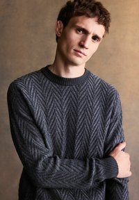 Grauer gestrickter Pullover mit Fischgrätenmuster, mit Rundhalsausschnitt und gerippten Bündchen, vor einem neutralen Hintergrund präsentiert.