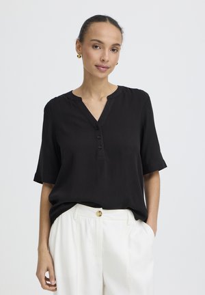 Mujer con blusa negra de manga corta con botones combinada con pantalones blancos de talle alto, posando frente a un fondo liso.