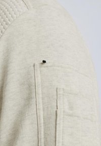 Sweatshirt beige clair avec un tissu texturé sur l'épaule, une poche poitrine et une petite étiquette noire près du bord de la poche.