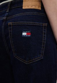 Σκούρο μπλε τζιν με πορτοκαλί ραφές. Διαθέτει πίσω τσέπη με μικρό κεντητό λογότυπο "TOMMY JEANS" σε κόκκινο, άσπρο και μπλε, καθώς και δερ μάτινη ετικέτα.