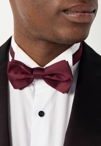 Un nœud papillon en satin bordeaux avec une texture lisse, porté sur une chemise blanche et une veste de smoking noire ; présente un bouton noir rond au niveau du col.