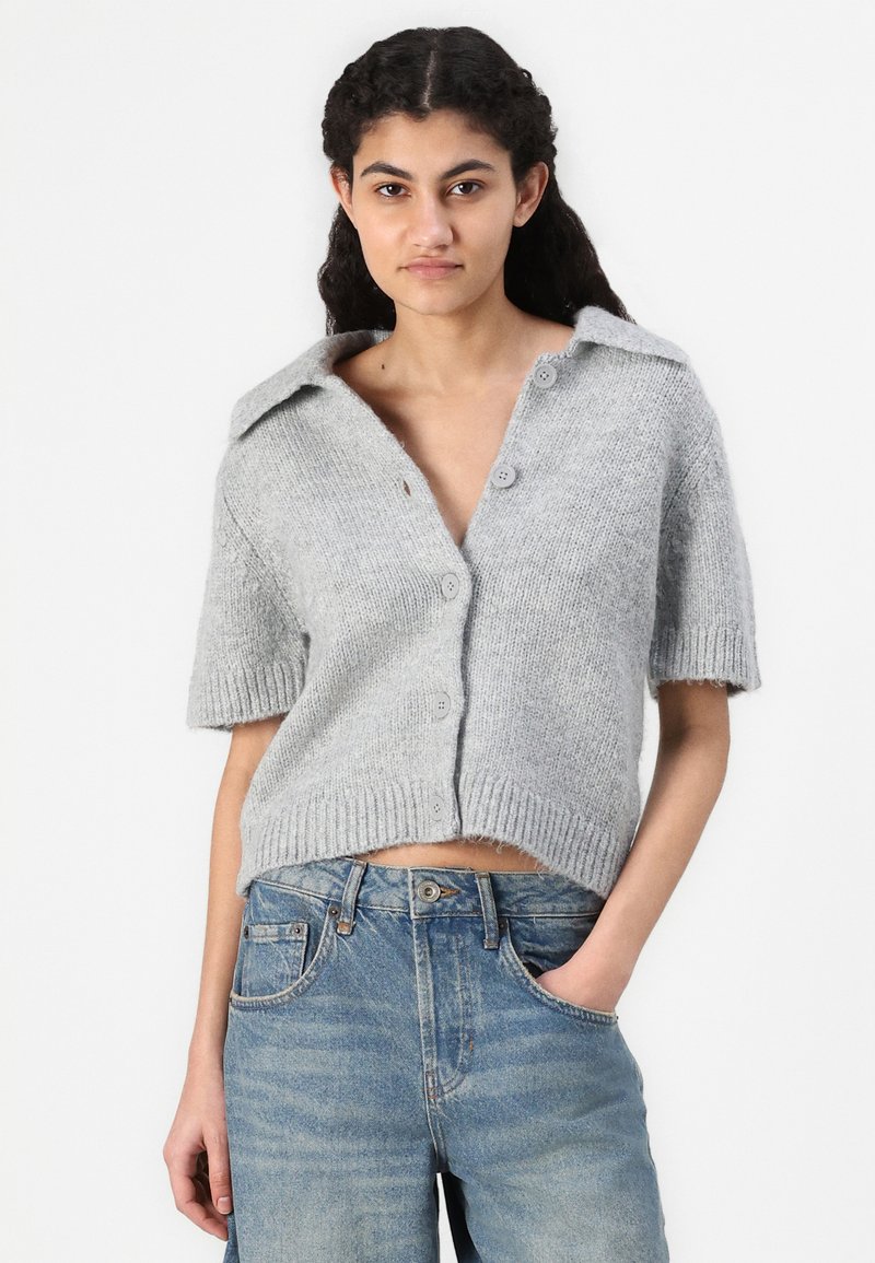 Gilet court en tricot gris à manches courtes avec un large col, fermeture à boutons, design court et ourlet côtelé. Porté sur un jean en denim.