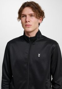 Svart zip-up träningsjacka i slät tyg, med hög krage och en vit chevron-logotyp på vänster bröst.