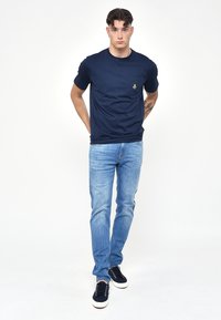 Mörkblå bomull T-shirt med en liten ficka och logotyp, kombinerad med ljusblå denimjeans och mörka sneakers på en enfärgad bakgrund.