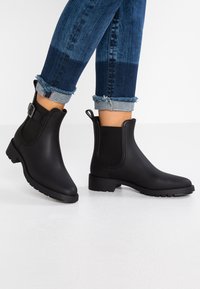 Bottes noires à la cheville en caoutchouc, avec des panneaux élastiques, un détail de boucle et une semelle texturée. Associées à un jean bleu retourné.