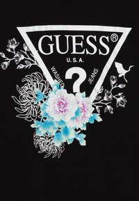 Musta puuvillainen t-paita, jossa suuri valkoinen kolmion muotoinen grafiikka, jossa tekstit "GUESS" ja "U.S.A.", sininen ja vaaleanpunainen kukkakirjonta sekä ääriviivakoristeluja.