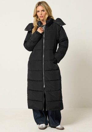 Femme portant un long manteau d'hiver matelassé noir avec une fermeture éclair à l'avant, debout avec une main dans la poche et l'autre près du col, fond neutre.