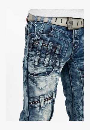 Pantalons cargo en denim bleu avec délavage, comportant plusieurs poches, des rayures noires et des détails de couture blanche contrastants.