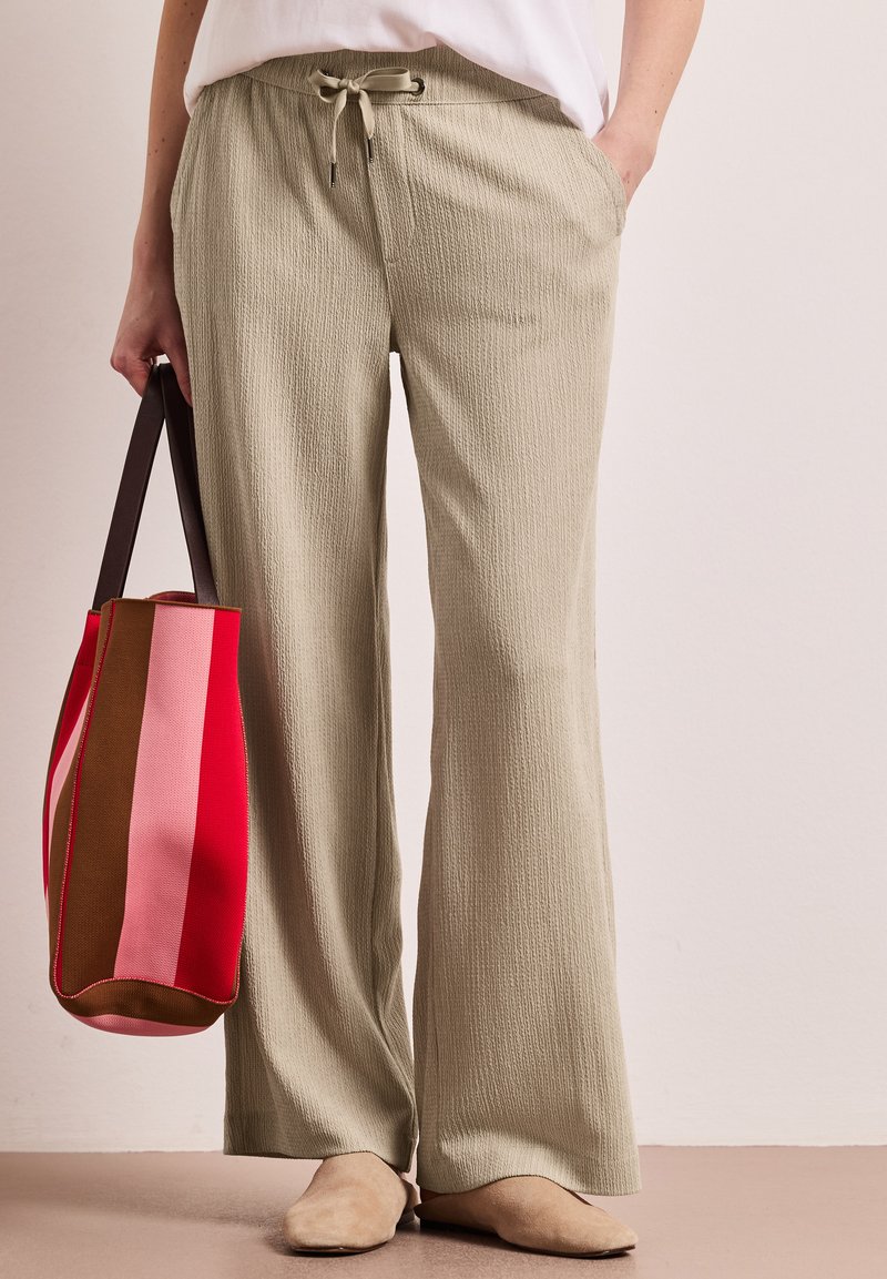 Kvinde iført beige, teksturerede bukser med vide ben og beige sko, der holder en lodretstribet taske i røde, pink og brune nuancer med mørke håndtag.