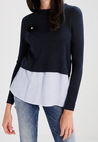 Marineblaues Strickpullover mit einem weißen gestreiften unteren Bereich und einer dekorativen schwarzen Blume mit einem Juwelenakzent auf der Brust. Lange Ärmel, runder Ausschnitt.