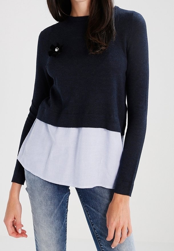 Marineblaues Strickpullover mit einem weißen gestreiften unteren Bereich und einer dekorativen schwarzen Blume mit einem Juwelenakzent auf der Brust. Lange Ärmel, runder Ausschnitt.