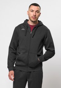 Fekete cipzáras kapucnis pulóver, puha anyagból készült, elülső zsebbel, állítható húzózsinóros kapucnival és kis fehér logóval a bal mellrészen.