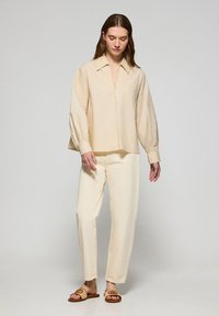 Blusa beige oversize con colletto e tasca frontale, abbinata a pantaloni larghi crema. Sandali comodi completano il look.