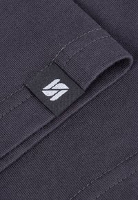 Dunkelgrauer Stoff mit gerippter Textur, ausgestattet mit einem gefalteten Rand und einem schwarzen Etikett mit einem weißen Logo an der unteren Naht.