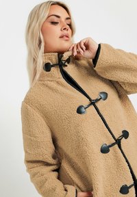 Beige fleece takki korkealla kauluksella, mustilla kiinnitysosilla ja teksturoidulla kankaalla. Suora vuori vetoketjun ja hihansuiden kohdalla.