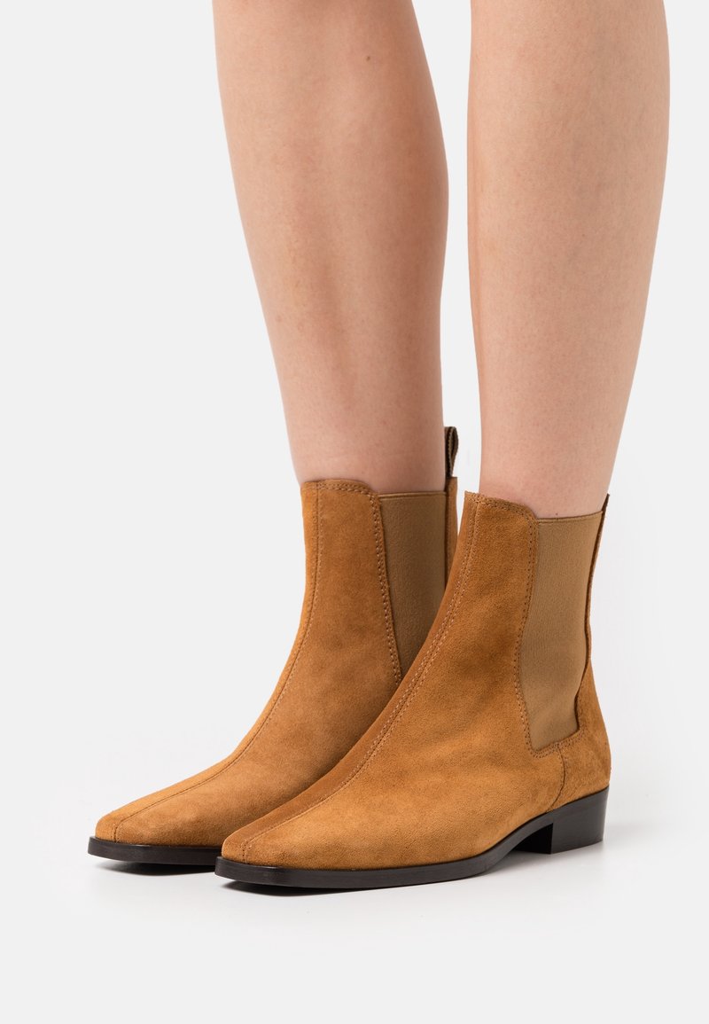 Bottes en daim beige montant à la cheville avec panneaux latéraux élastiques et petits talons noirs portées sur jambes nues sur fond blanc.