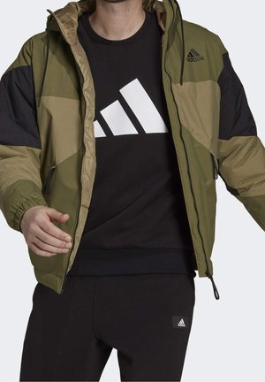 Homme portant une veste Adidas verte et noire dézippée sur un sweat-shirt Adidas noir et un pantalon noir, debout devant un fond blanc.
