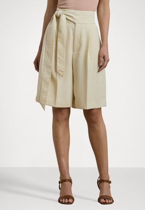Beige shorts med høj talje med plisseringer og et langt stofbælte bundet på venstre side, båret med brune sandaler med ankelrem.
