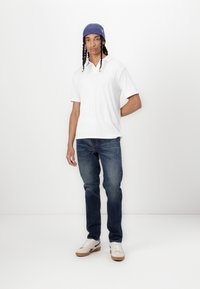 Camisa polo blanca de manga corta y canalé, pantalones vaqueros de color azul oscuro y zapatillas beige con detalles oscuros. La persona lleva un gorro azul.