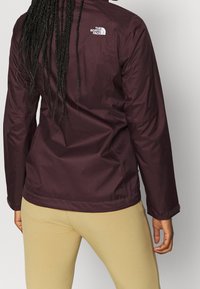 Bordeauxrode waterdichte jas met een hoge kraag, lange mouwen en een logo op de bovenrug. Het materiaal lijkt glad en licht van gewicht.