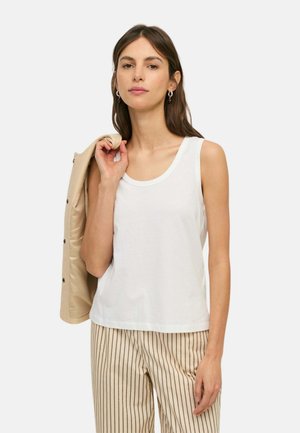 Mujer con top blanco sin mangas y pantalones beige con rayas, sosteniendo una chaqueta beige sobre su hombro sobre un fondo liso.