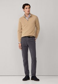 Suéter beige con cremallera, camisa de color azul claro debajo con cuello, pantalones chinos grises y zapatos de ante oscuros. Diseño simple y ajustado con líneas horizontales.