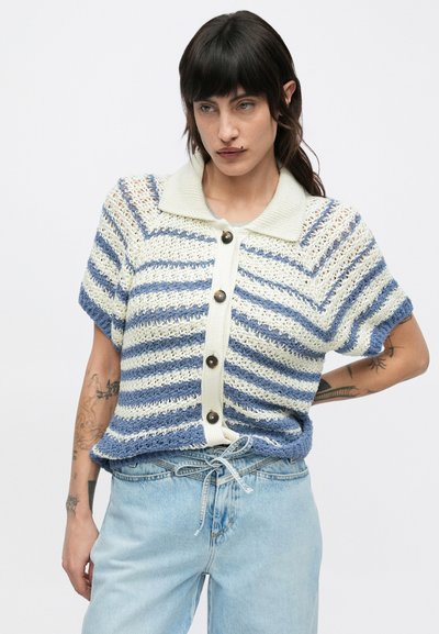 Γυναίκα με σκούρα μαλλιά που φοράει μπλε και λευκή ριγέ πλεκτή ζακέτα με κοντά μανίκια και ανοιχτόχρωμο τζιν με δεμένη ζώνη, στέκεται με το ένα χέρι στη μέση.