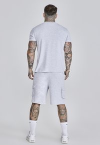 T-shirt en coton gris et short assorti avec poches cargo, portés avec des chaussettes blanches et des baskets. Tatouages visibles sur les bras et les jambes.
