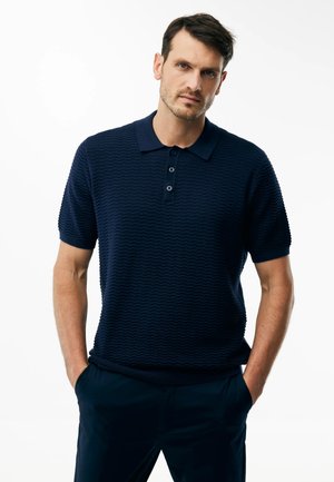 Mann mit kurzen dunklen Haaren, der ein marineblaues, strukturiertes Poloshirt mit kurzen Ärmeln und marineblaue Hosen trägt, steht mit den Händen in den Taschen.
