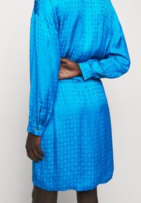 Blå satin klänning med långa ärmar, med ett subtilt geometriskt mönster. Midjan är elastisk och har guldknappar vid ärmsluten.