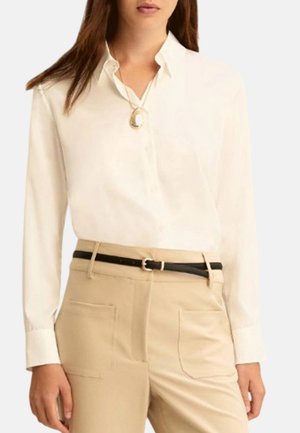 Donna che indossa una camicia bianca con bottoni, pantaloni beige con tasche frontali, cintura nera e collana con ciondolo dorato.