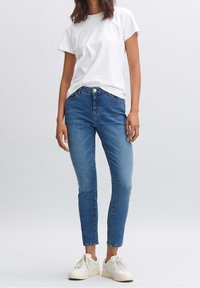 Žena v bílém tričku, modrých skinny džínách s vysokým pasem a bílých teniskách stojí proti čistému šedému pozadí.