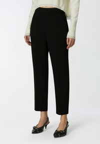 Crne krojene hlače s glatkom teksturom, suženim oblikom i strukom srednje visine, uparenima s crnim cipelama na visoku petu s uskim nosom i kopčama.