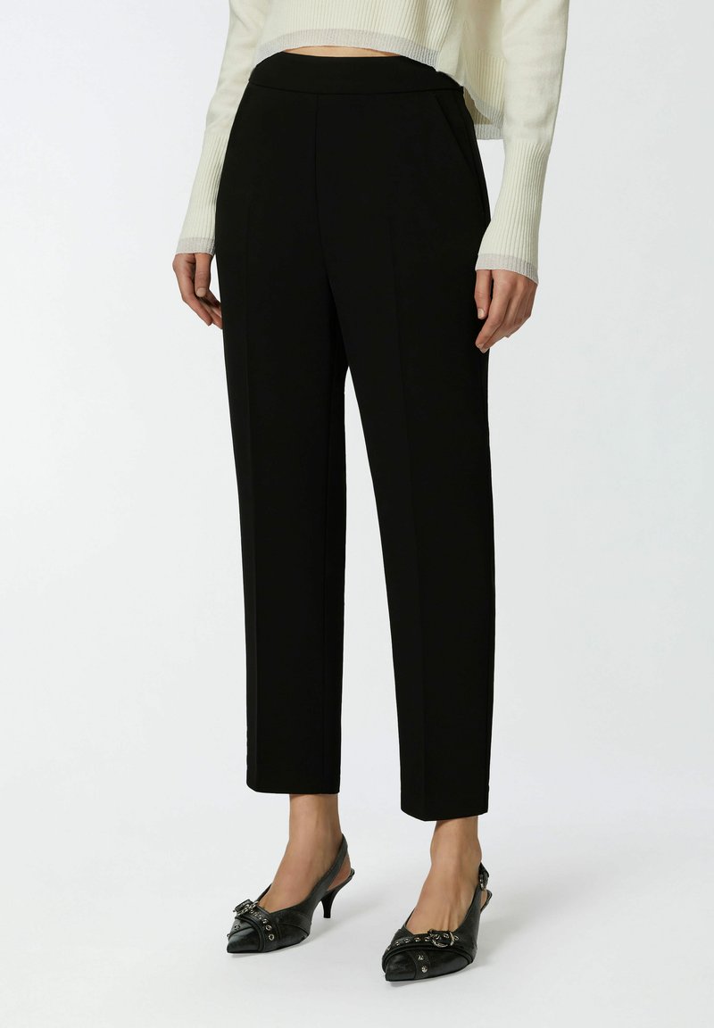 Crne krojene hlače s glatkom teksturom, suženim oblikom i strukom srednje visine, uparenima s crnim cipelama na visoku petu s uskim nosom i kopčama.