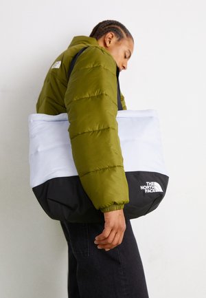 Person mit grüner Steppjacke trägt große schwarz-weiße The North Face Tragetasche über der Schulter vor einfarbigem Hintergrund.