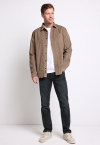 Brązowa koszula z guzikami, z kieszonką na piersi, noszona na białej koszulce. Połączona z ciemnoniebieskimi dżinsami i jasnymi trampkami, stojąca na jednolitym tle.