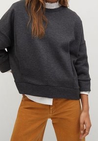 Pull gris avec col rond, épaules tombantes et coupe ample, associé à un pantalon beige. Texture lisse, détails minimaux.